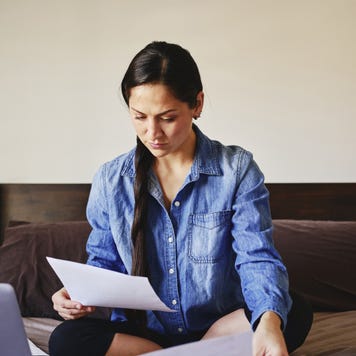 A young woman is sitting cross-legged on a couch and reviewing some financial forms.