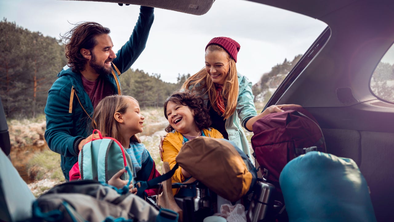 Two smiling adults and two smiling children taking backpacks out of an open trunk