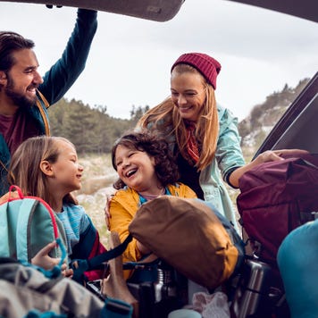 Two smiling adults and two smiling children taking backpacks out of an open trunk