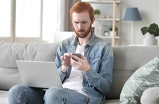 Red haired man sits on white sofa with laptop on knees and scrolls phone, looking pensive