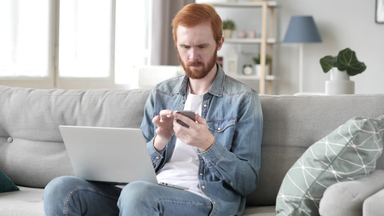 Red haired man sits on white sofa with laptop on knees and scrolls phone, looking pensive