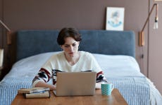 Woman sitting in front of bed at a wooden table with laptop open and mug nearby.