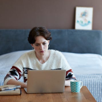 Woman sitting in front of bed at a wooden table with laptop open and mug nearby.