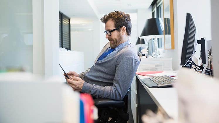 Male in grey cardigan browsing smartphone in the office | Hero Images/Getty Images