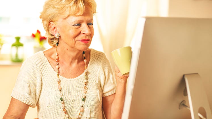 Senior woman holding cup of drink while on the computer  © iStock