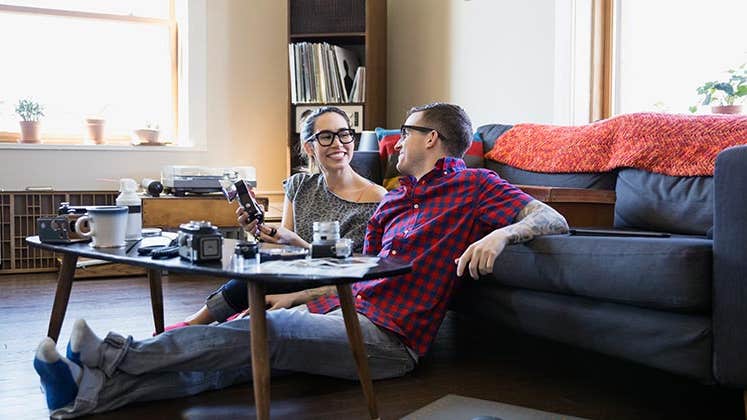 Smiling millennial couple sitting on living room floor | Hero Images/Getty Images