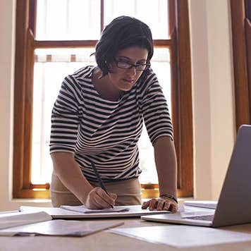 Woman leaning over and writing on paper | PeopleImages/DigitalVision/Getty Images