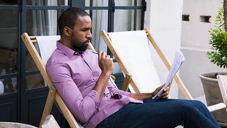 Man in purple shirt reading paperwork in patio chair | PhotoAlto/Sigrid Olsson/Getty Images