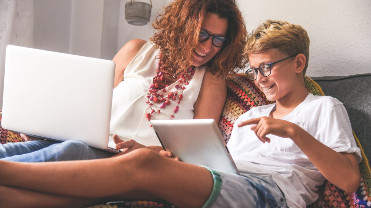 Mother and pre-teen son sit together on bed working on laptop and tablet