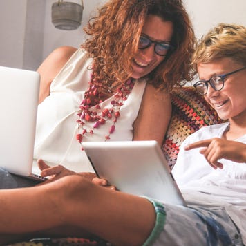 Mother and pre-teen son sit together on bed working on laptop and tablet