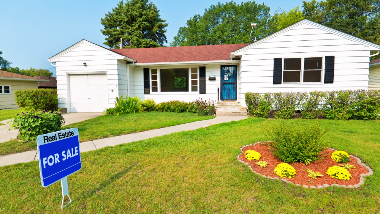 white suburban house with green front yard and landscaping, for sale sign out front