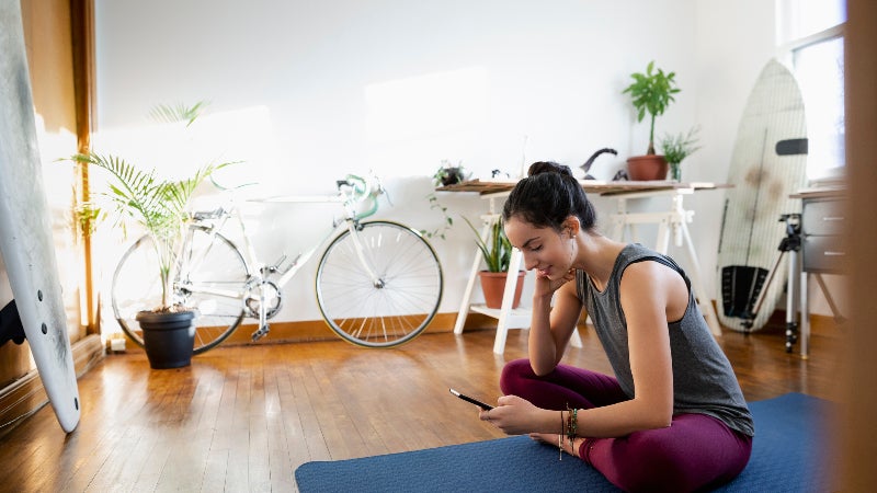 Young woman with smart phone on yoga mat in apartment