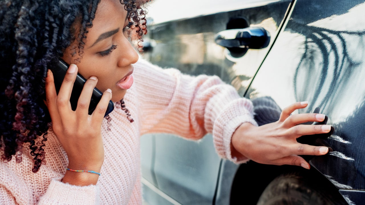 Younger black lady looking at damage to her car and talking on her phone to her insurance provider