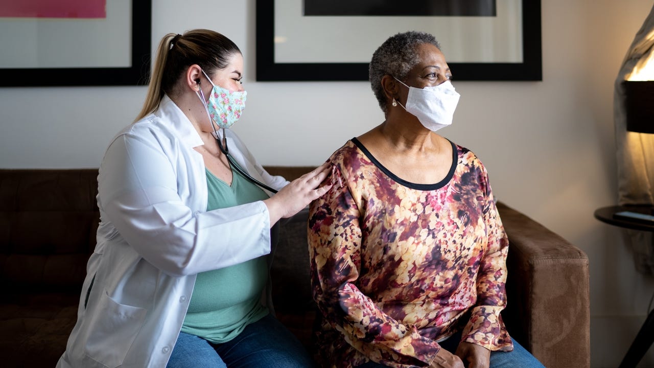 Doctor using stethoscope listening to senior patient breathing at her house - using face mask