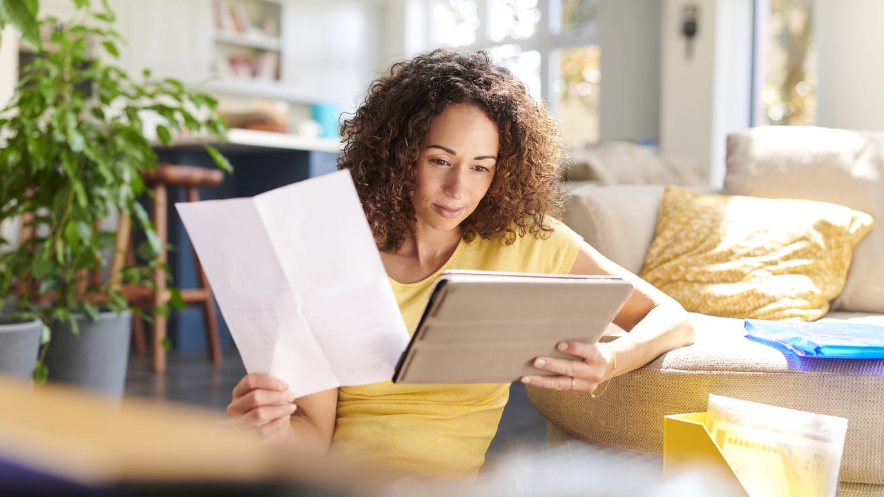 A woman in her living room holding a tablet in one hand and a piece of paper in the other. She is checking her home finances online.