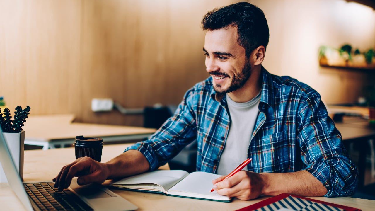 man at desk with laptop writing in notebook