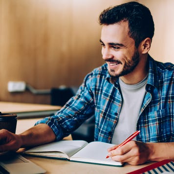 man at desk with laptop writing in notebook