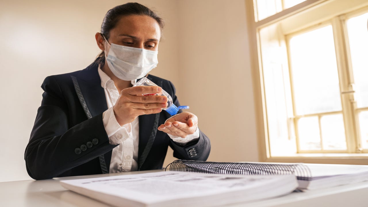 A woman with a facemask applies hand sanitizer while looking over a binder of information.