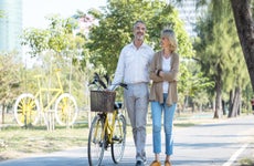 A senior couple walking together on a sunny day. The man is pushing a bicycle with a basket on it.