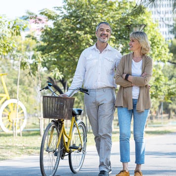 A senior couple walking together on a sunny day. The man is pushing a bicycle with a basket on it.