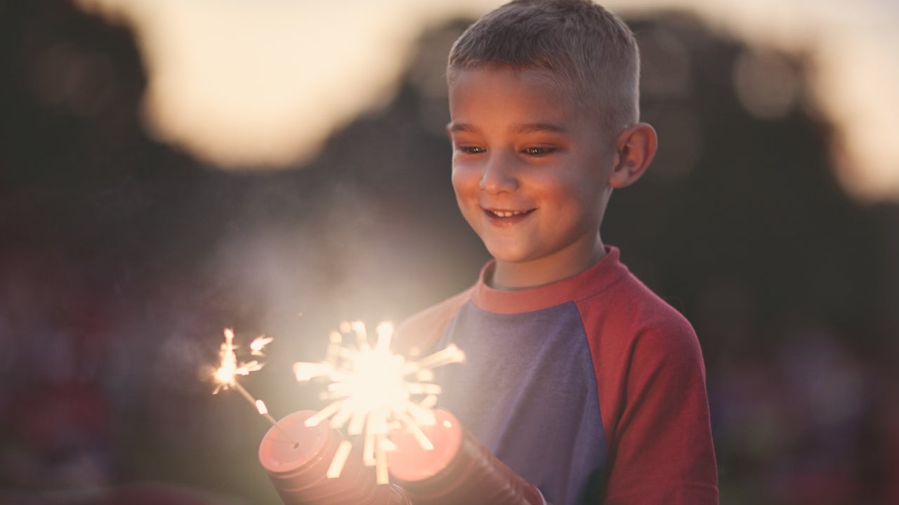 A boy holds two sparklers and stares at them in wonder (his hands are protected by two red, plastic cups where holes have been poked in them to hold the sparkler sticks)