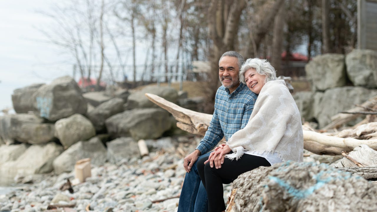 An older couple sitting together on a rocky beach.