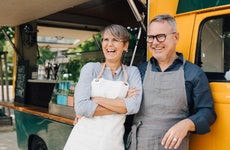 An older White couple stands with aprons in front of a food truck