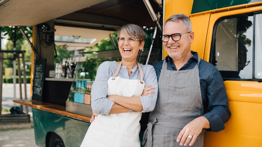 An older White couple stands with aprons in front of a food truck
