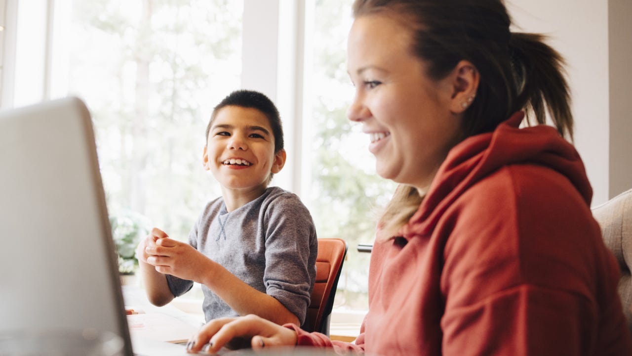 A mother sits at a computer while her special needs child sits next to her; they are laughing.