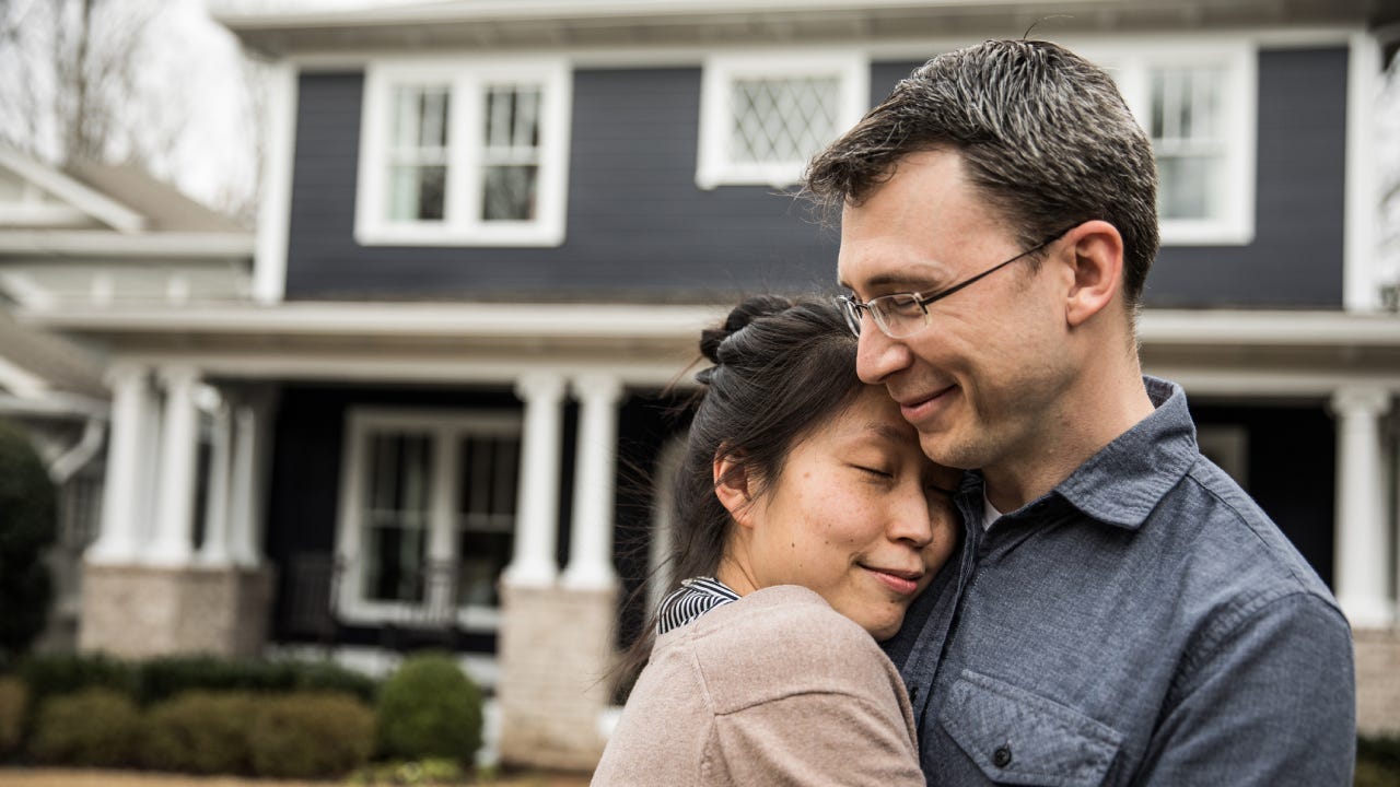 A man and woman embrace in front of their home.