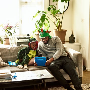 A father and his son, dressed up like dragons, do a learning exercise in the living room.