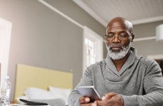 An older black man sits at his table with his phone and a list of finance reports to review.
