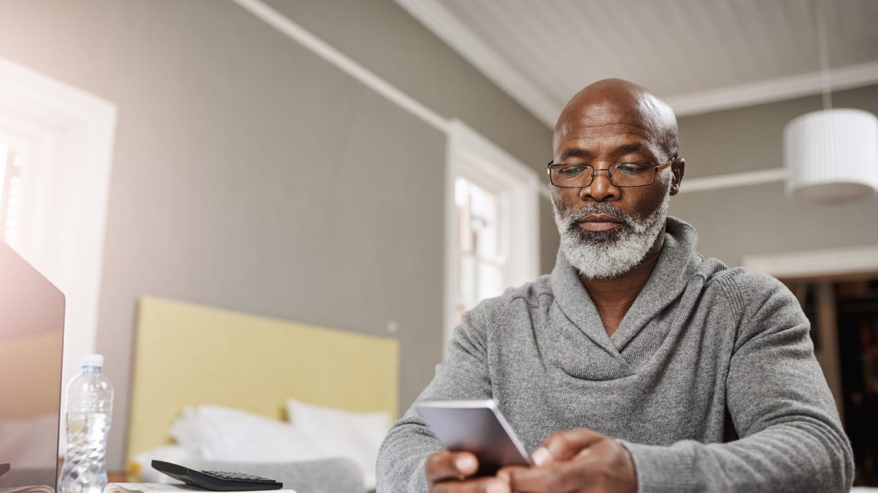 An older black man sits at his table with his phone and a list of finance reports to review.