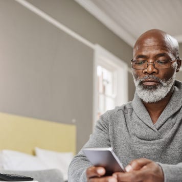 An older black man sits at his table with his phone and a list of finance reports to review.