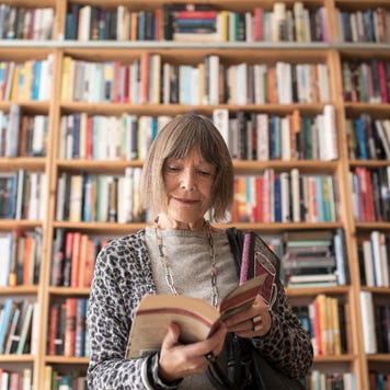 An old white woman flips through a book in front of a bookshelf