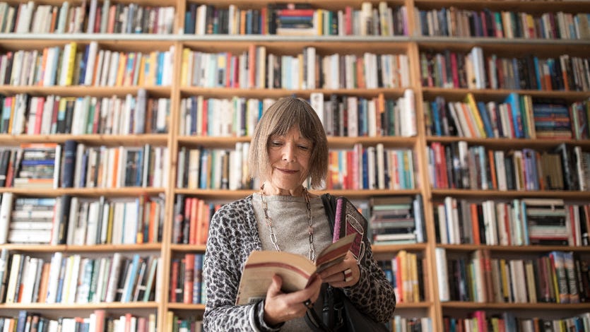 An old white woman flips through a book in front of a bookshelf