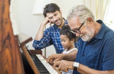 An old man sits with his son and granddaughter and plays on the piano.