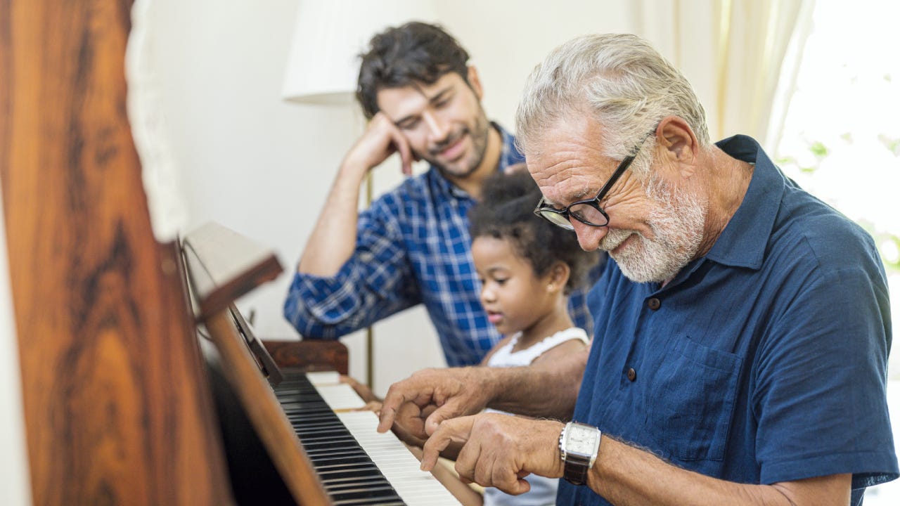 An old man sits with his son and granddaughter and plays on the piano.
