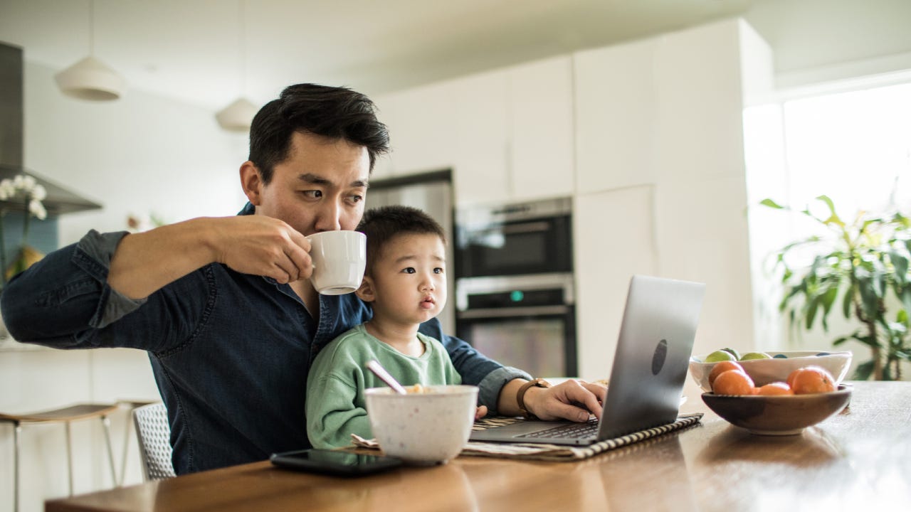 A father sits with his son in front of the laptop while sipping his coffee.