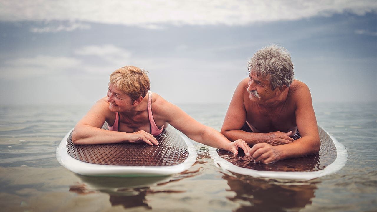 An older couple on surf boards holding hands on some calm water.