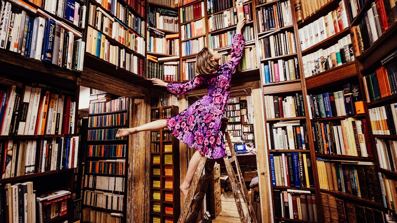 A woman stands on a ladder in a bookstore to reach the highest shelf