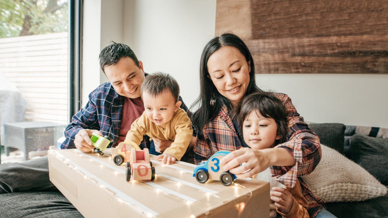 A family sits with their kids, playing with toy cars on a cardboard box made to look like a racetrack.