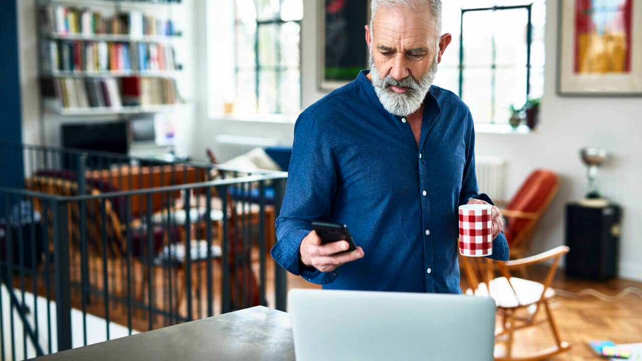 Man in his 50s in apartment, texting, with laptop on work surface, serious expression on face, planning for the day ahead, drinking coffee