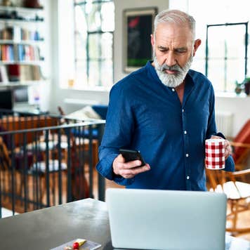 Man in his 50s in apartment, texting, with laptop on work surface, serious expression on face, planning for the day ahead, drinking coffee