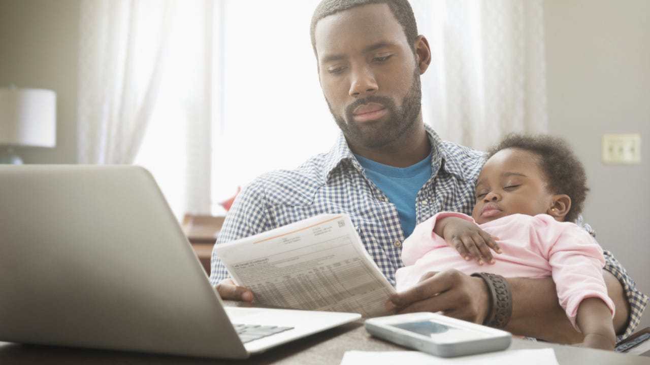 A single father sits with their child in one arm and a financial record in the other, reviewing finances.