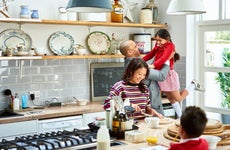 A father holds his daughter up in the air while mom is making lunches and smiling in the kitchen.
