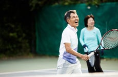 An older Asian man holds a tennis racket on a court and shouts in joy after scoring a point.