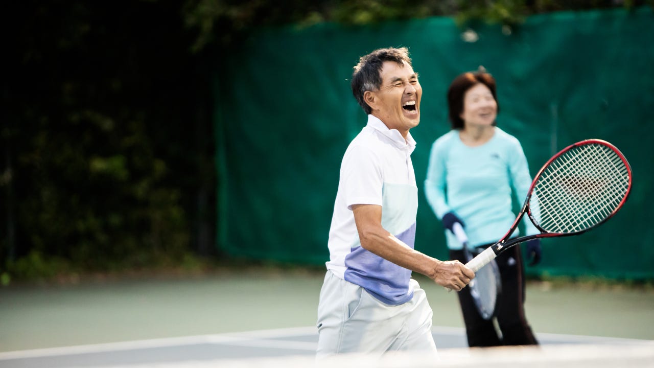 An older Asian man holds a tennis racket on a court and shouts in joy after scoring a point.