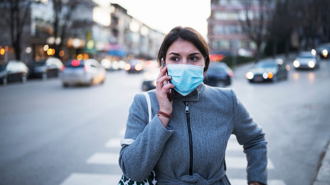 A women wearing a facemask talks on the phone while crossing the street