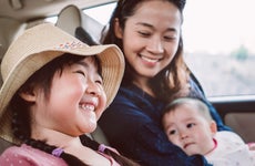 Asian woman sits in the back seat of a car with her infant child in her arms. Her daughter sits next to her, smiling and wearing a floppy straw hat.